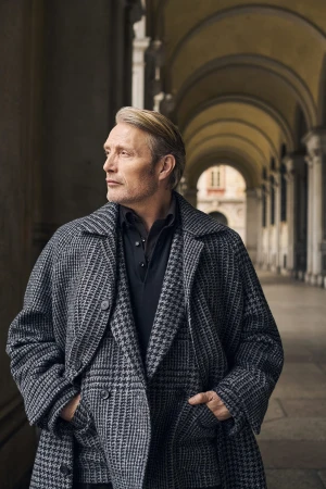 Mads Mikkelsen wearing a grey houndstooth coat and dark shirt, standing in an arched colonnade.