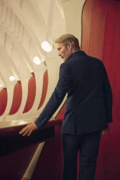 Mads Mikkelsen wearing a dark suit standing in an architectural interior with curved white ribs and red panels for Zegna Fall 2025 campaign.