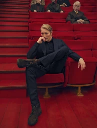 Mads Mikkelsen seated in a red theatre chair wearing a dark suit for the Zegna Fall 2025 campaign
