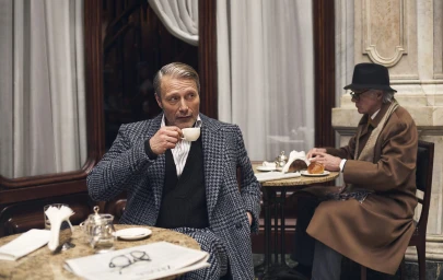 Mads Mikkelsen drinking from a small white cup in a houndstooth coat at a cafe table for Zegna Fall 2025 campaign.