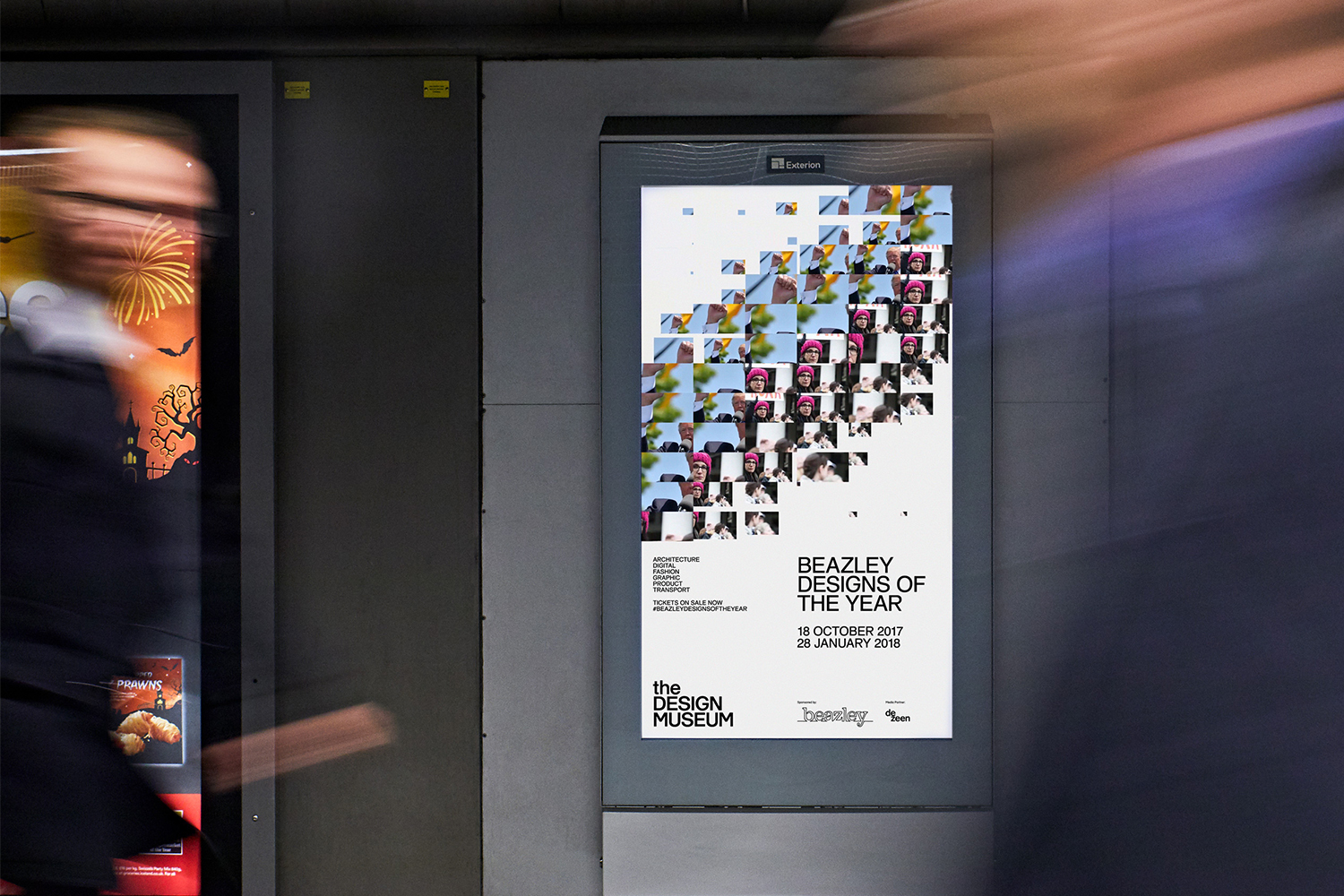 Digital poster displaying Beazley Designs of the Year exhibition on a public display panel with blurred commuters in motion foreground.