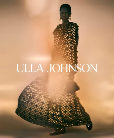 Model in patterned dark dress with gold earrings standing against a warm glowing backdrop for Ulla Johnson campaign