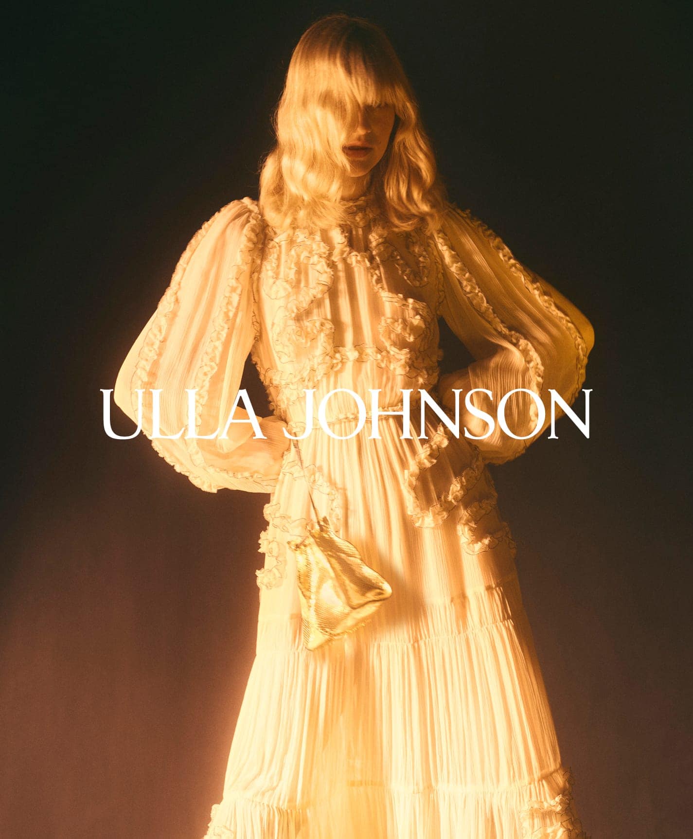 Blonde model in a white ruffled tiered dress with voluminous long sleeves holding a gold pouch bag against a dark background