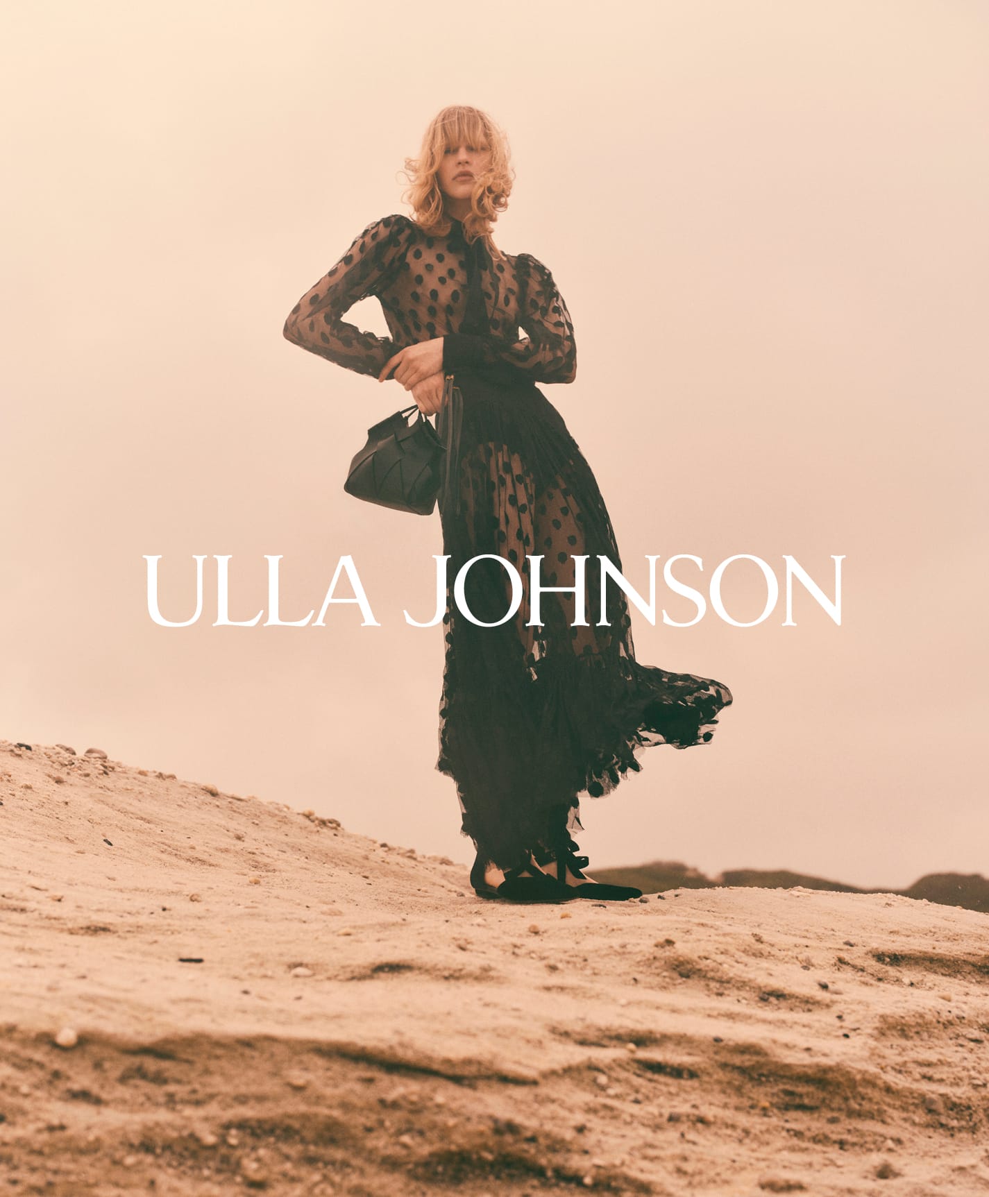 Model standing on a sandy hill wearing a black sheer dotted dress and holding a handbag in the Ulla Johnson Fall 2025 campaign.