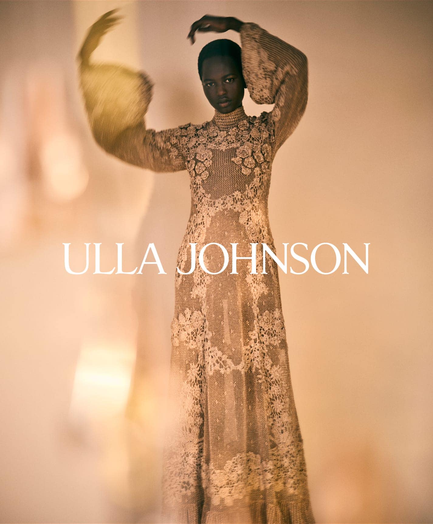 Model in intricate patterned lace long-sleeve dress raising her hands in front of a neutral gradient background.