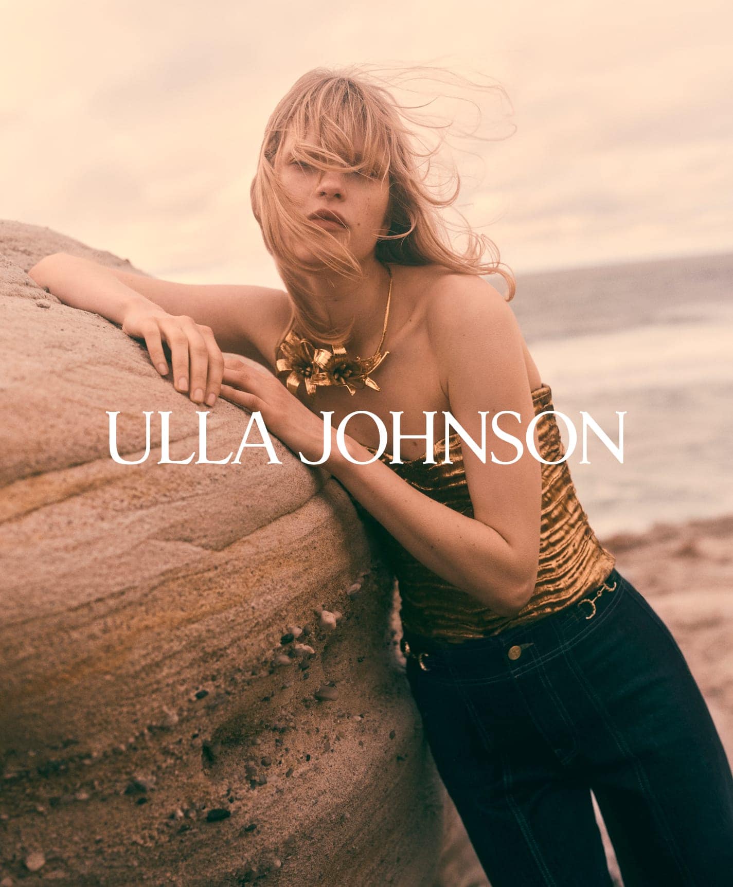 Model leaning against a large rock by the ocean wearing a gold top and dark trousers for Ulla Johnson Fall 2025 ad campaign.