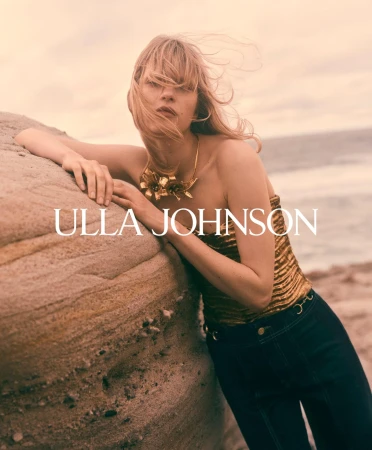 Model leaning against a large rock by the ocean wearing a gold top and dark trousers for Ulla Johnson Fall 2025 ad campaign.