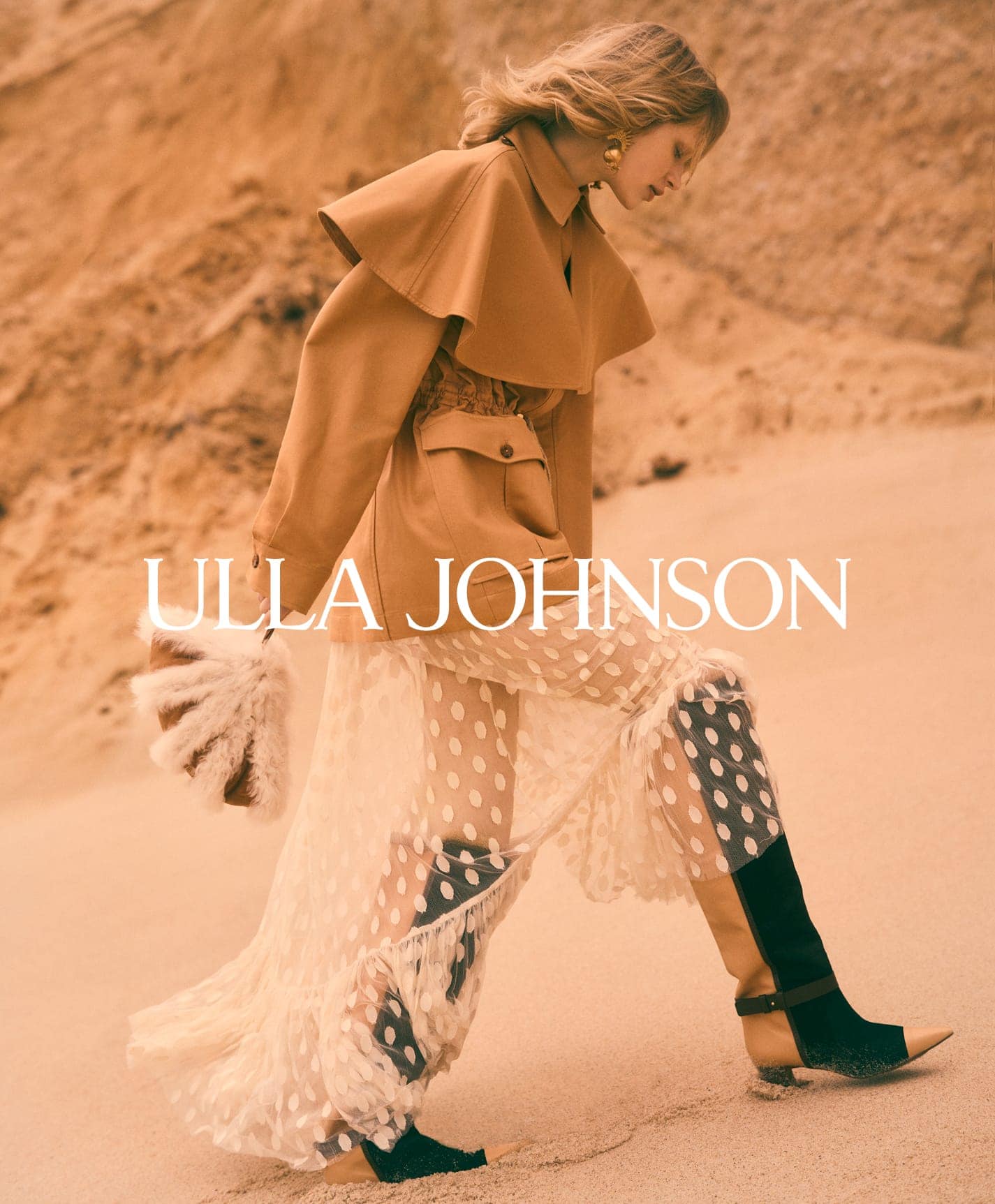 Model wearing a tan cape jacket and polka dot sheer skirt walking outdoors against a sandy earth backdrop