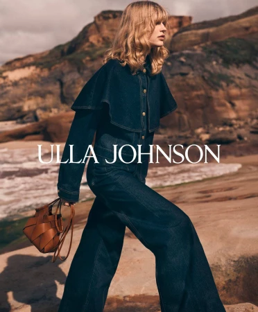Model in dark denim jacket and matching trousers holding a brown leather handbag on a coastal beach background