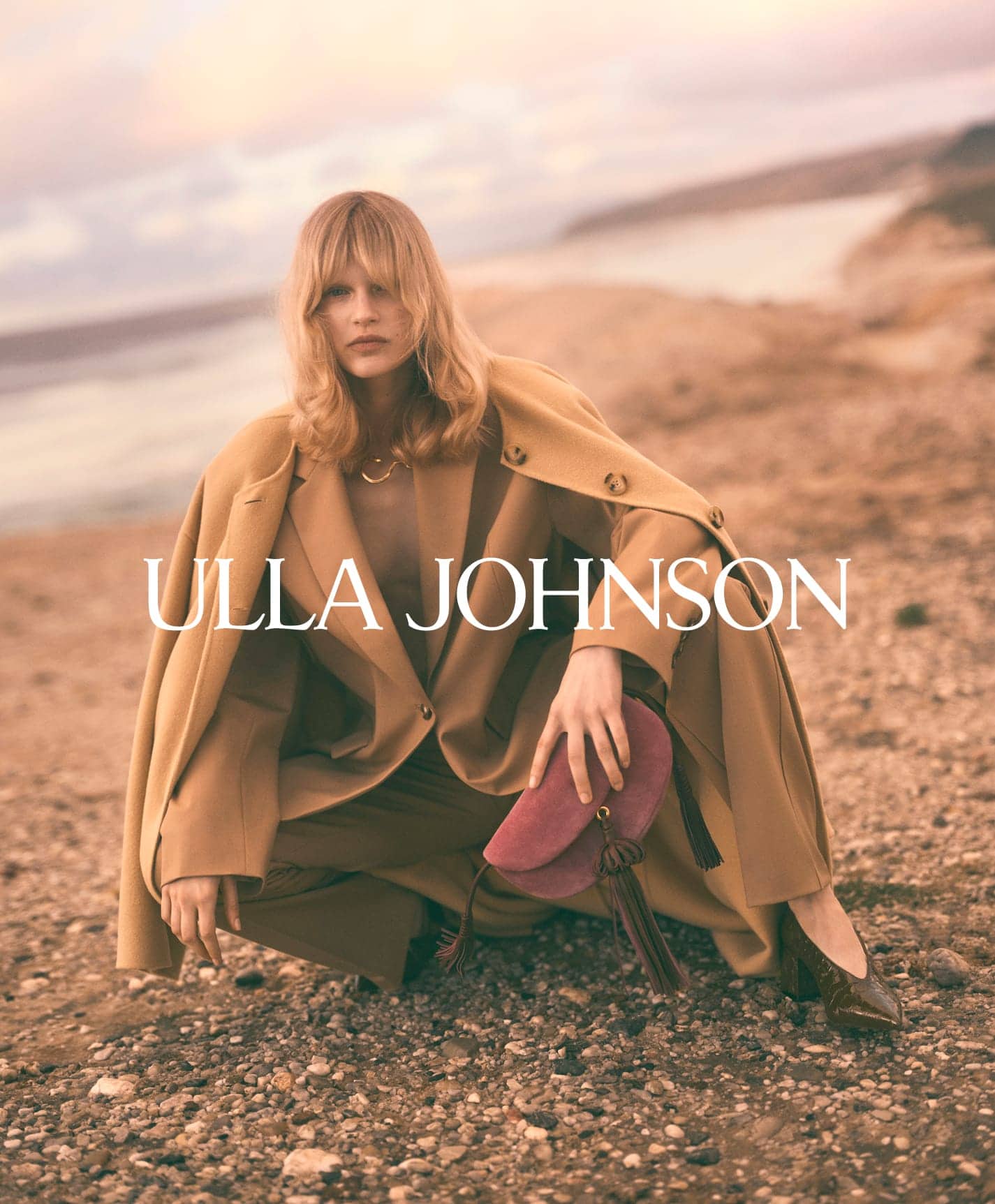 Model crouching on a rocky coastal shore wearing a tan coat and holding a pink suede handbag for Ulla Johnson.