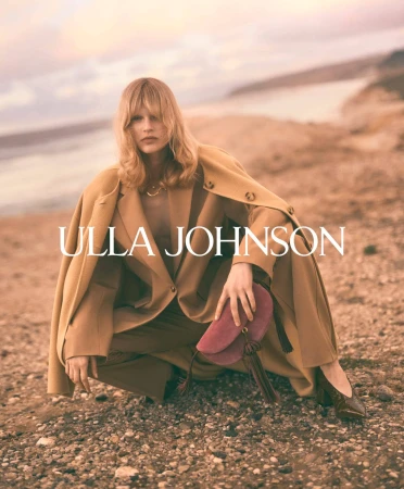 Model crouching on a rocky coastal shore wearing a tan coat and holding a pink suede handbag for Ulla Johnson.