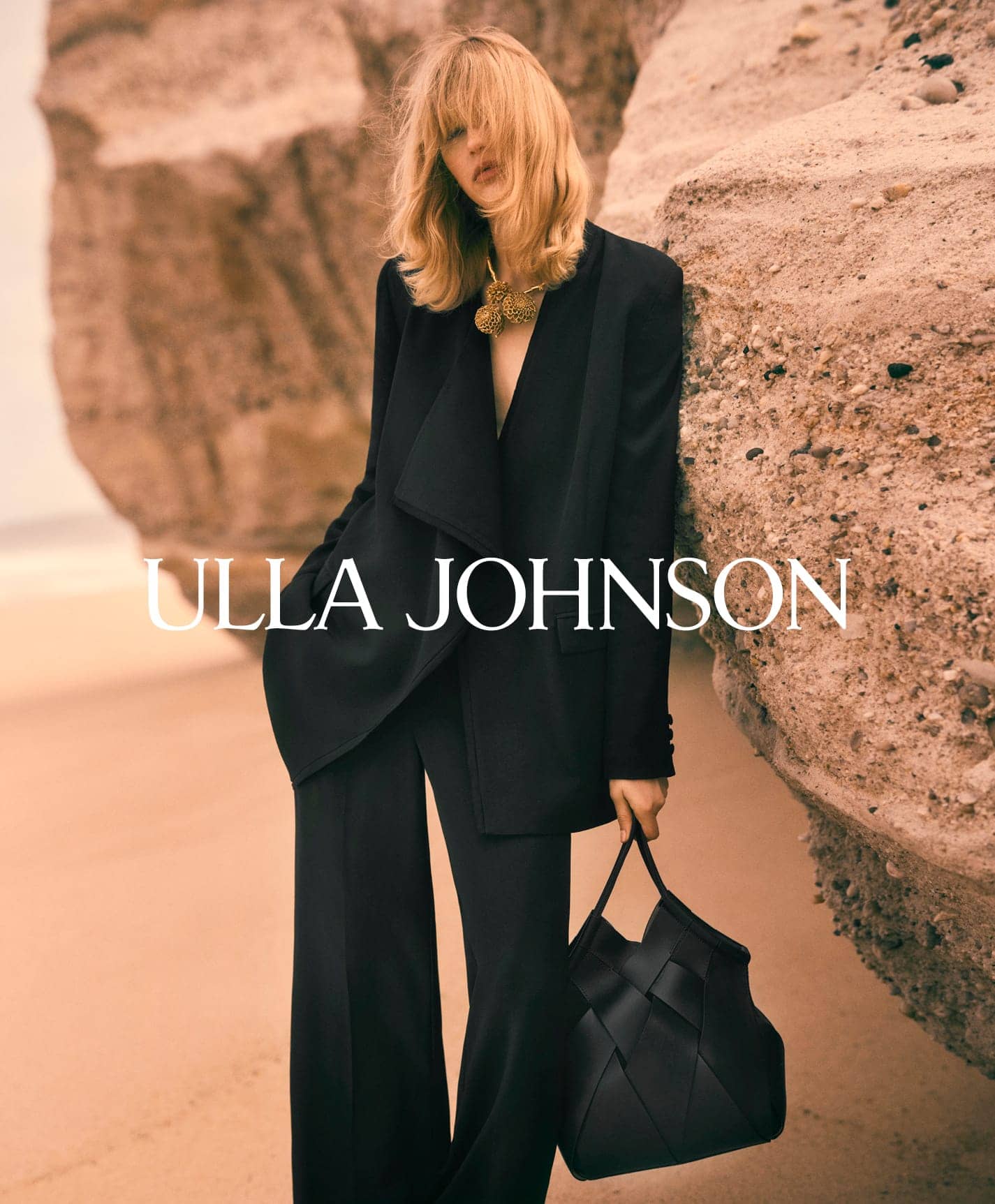 Model in black suit leaning against a rocky sandy cliff holding a black handbag