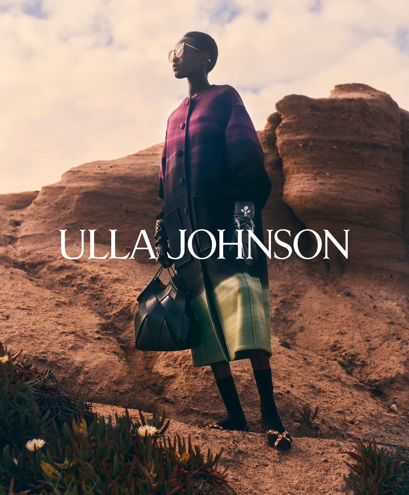 Model wearing purple gradient coat and black handbag standing outdoors on a rocky terrain under a cloudy sky.