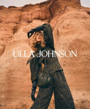 Model posing against a rugged earthen rock cliff wearing a patterned dark green top and matching trousers with leather gloves.
