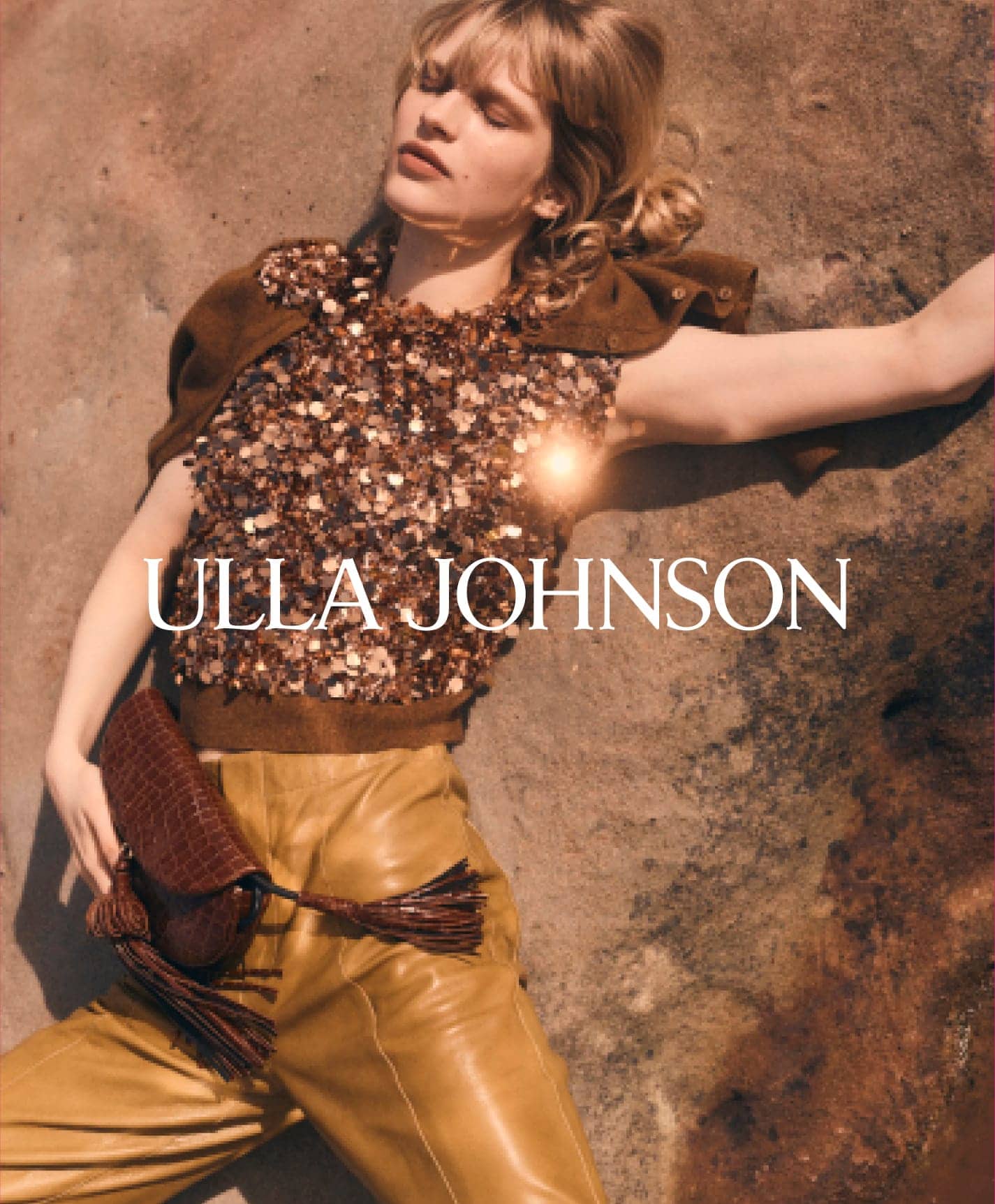 Model lying against a rock wearing a sequined top and tan leather pants holding a brown fringed clutch