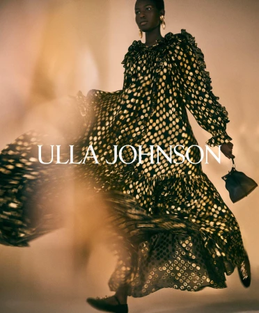 Model in a black dress with gold polka dots wearing gold earrings and holding a handbag for Ulla Johnson