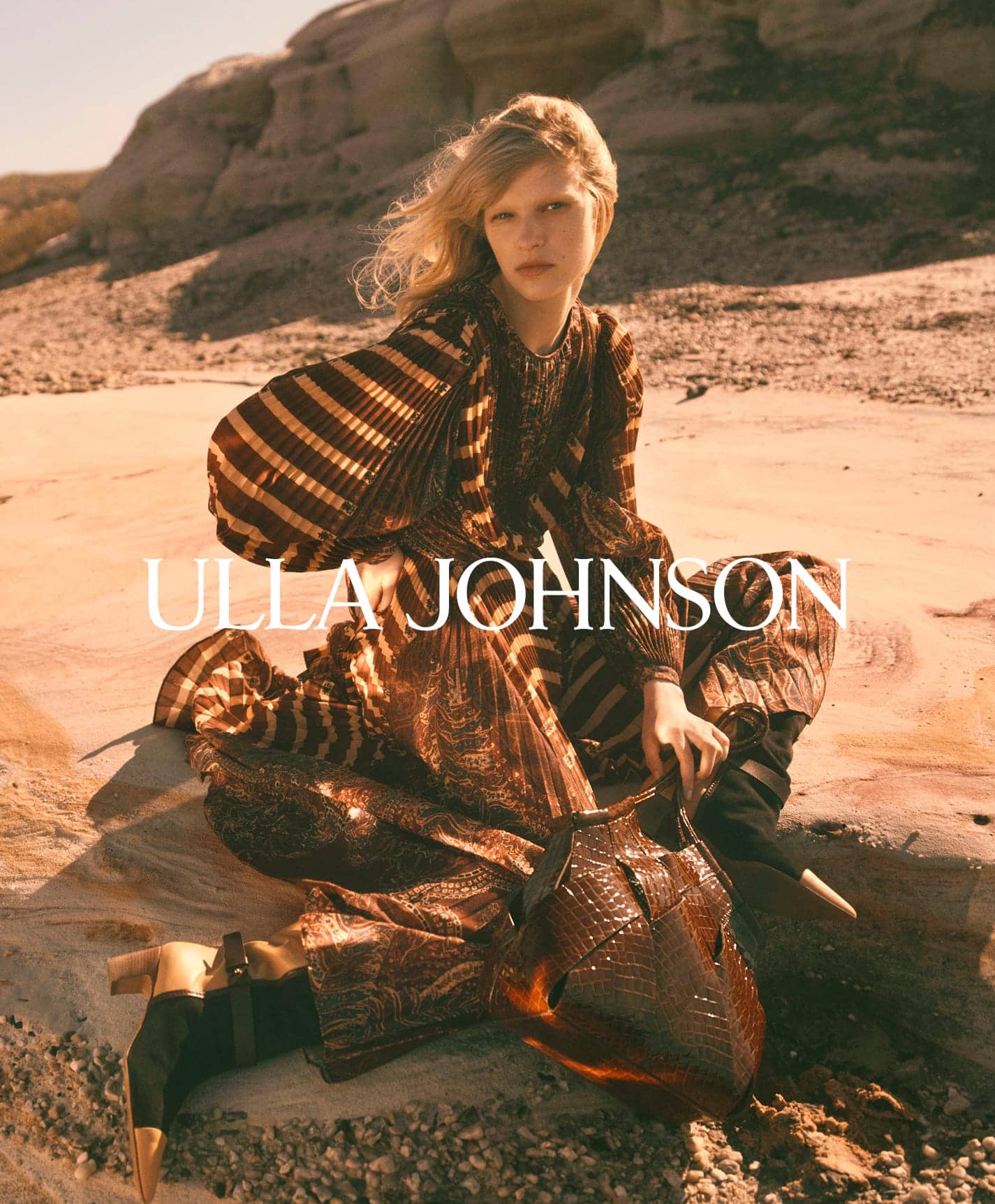 Model wearing a patterned brown dress and holding a woven leather bag outdoors on sandy terrain for Ulla Johnson.