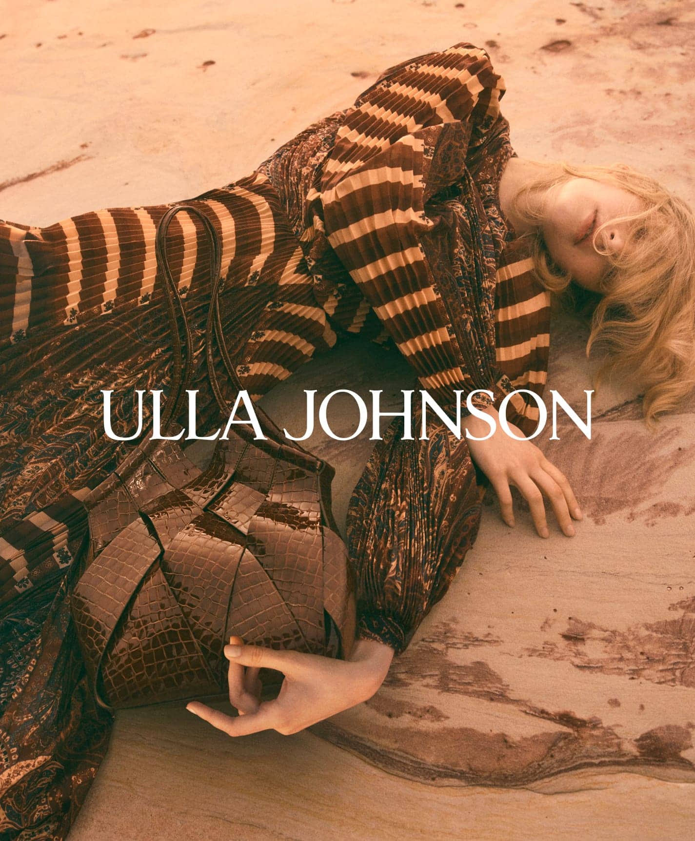 Model lying on sandy ground wearing a pleated brown patterned dress and holding a dark embossed leather handbag