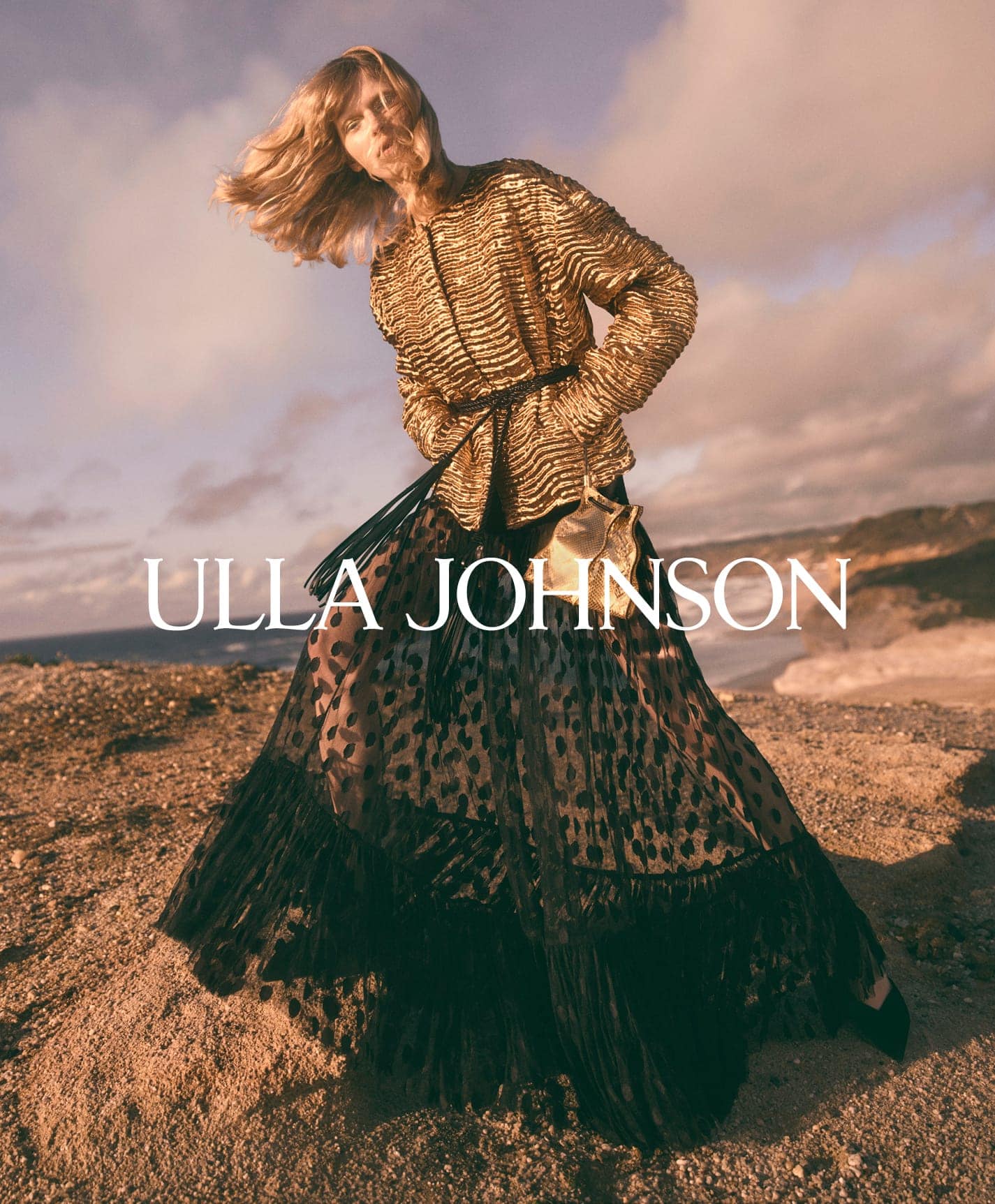 Model in a textured gold jacket and dark skirt standing on an outdoor coastal terrain at sunset