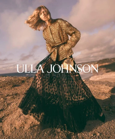 Model in a textured gold jacket and dark skirt standing on an outdoor coastal terrain at sunset