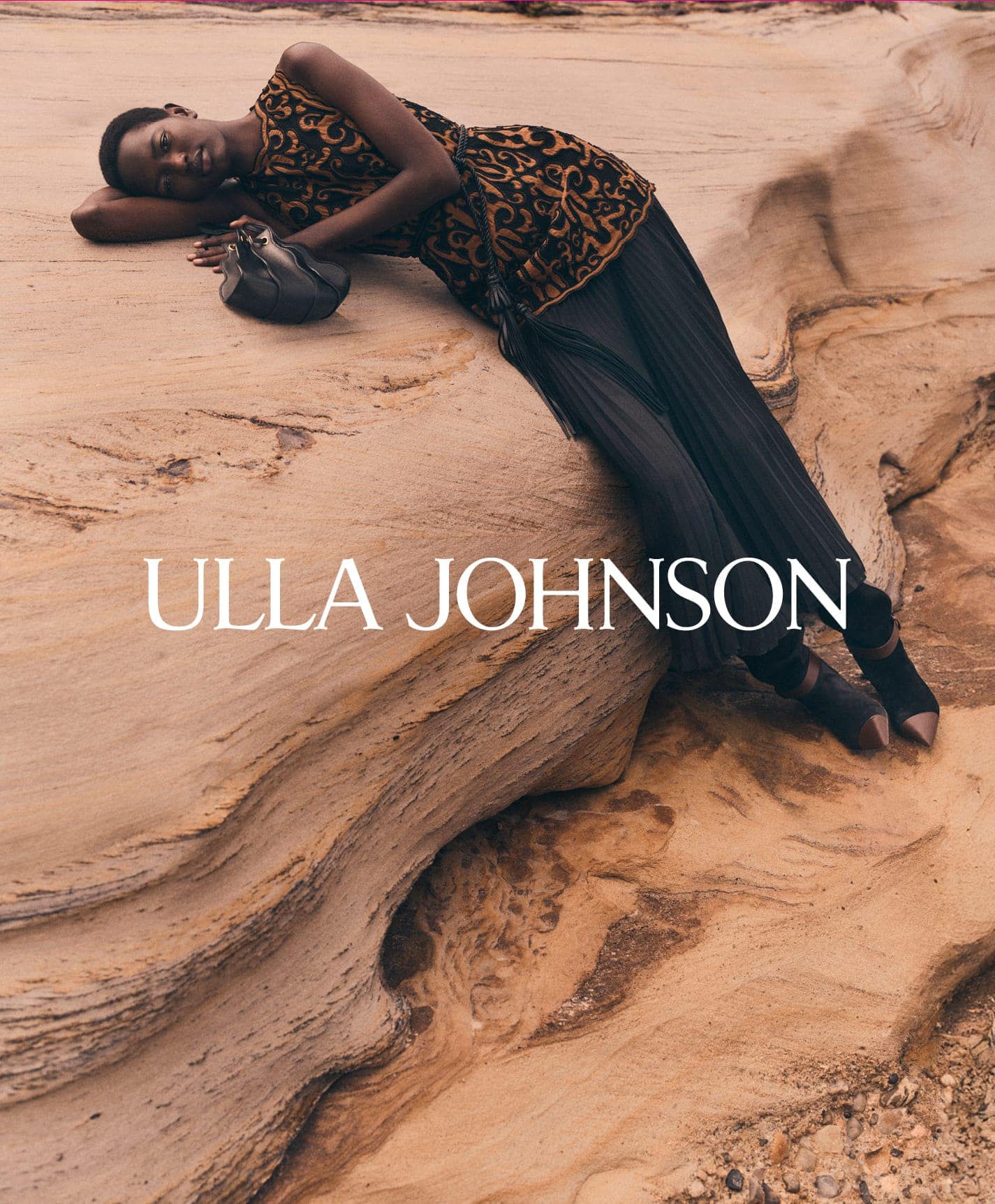 Model reclining on weathered rock formation wearing patterned top and pleated skirt for Ulla Johnson campaign