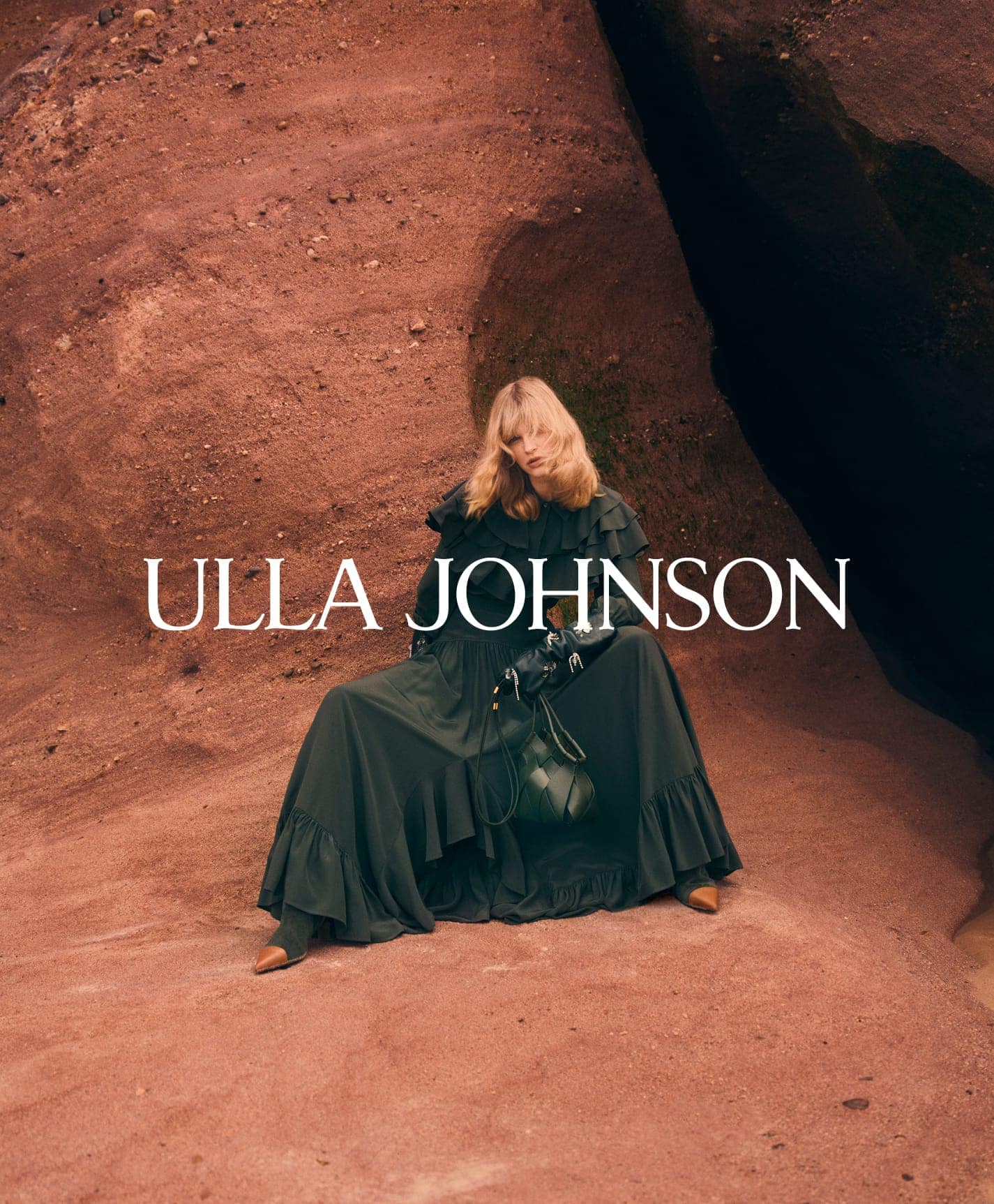 Model wearing a dark ruffle dress seated against a rugged red earth rock formation