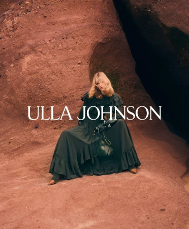 Model wearing a dark ruffle dress seated against a rugged red earth rock formation