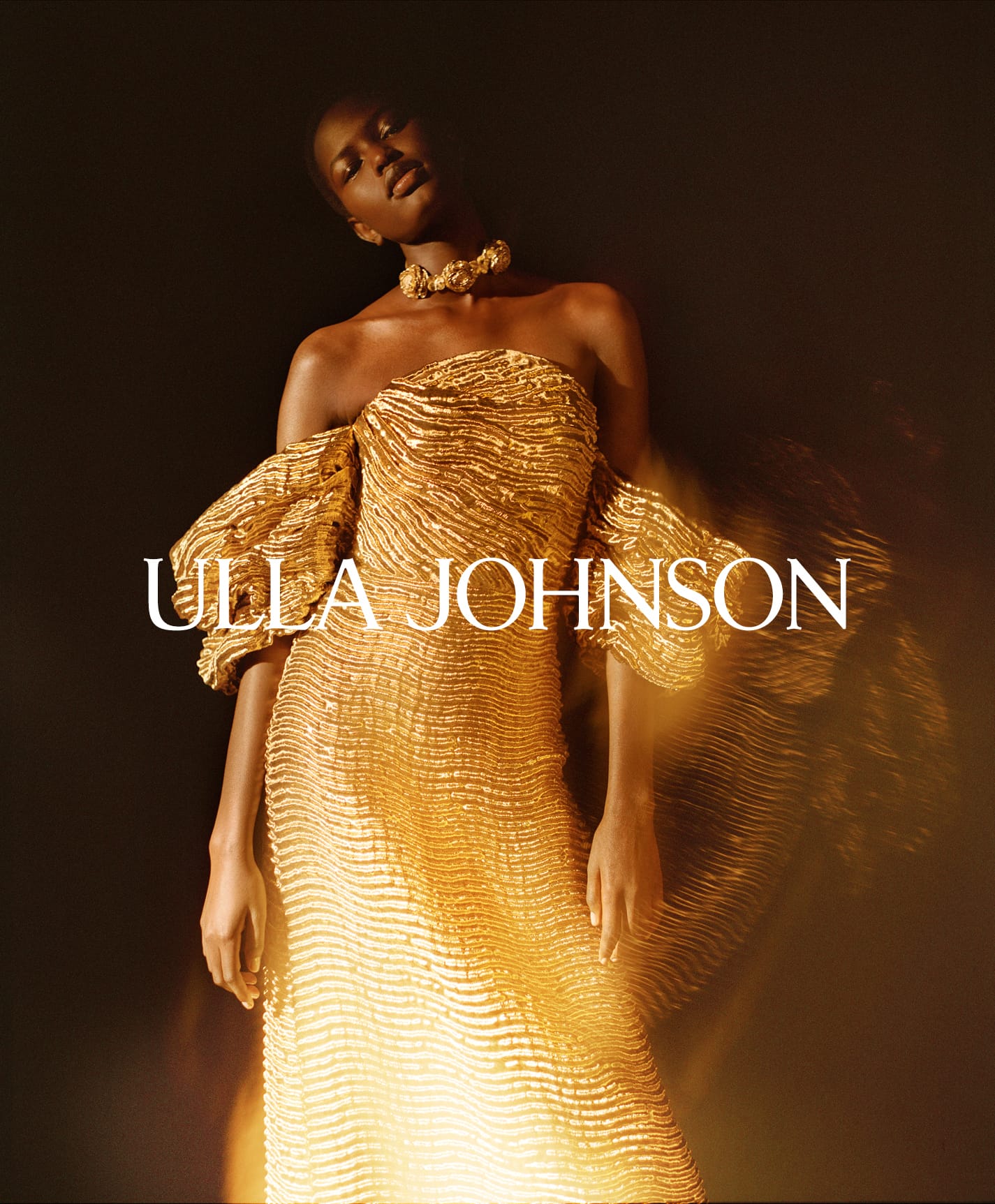 Model wearing a textured gold strapless gown with sculptural drop sleeves and a matching statement collar against a dark background.