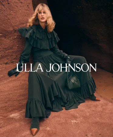 Model seated against a textured reddish rock formation wearing a ruffled dark green dress and holding a dark handbag.