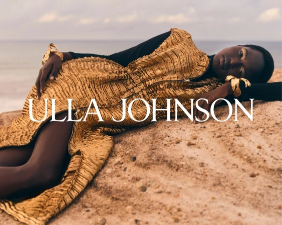 Model wearing a textured gold dress and gold cuffs lying on sand by the ocean