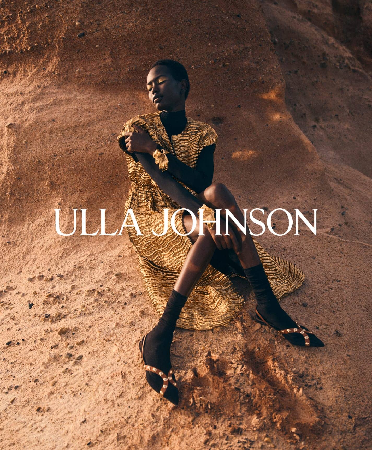 Model wearing a gold textured dress and dark socks seated against a textured rocky cliff background
