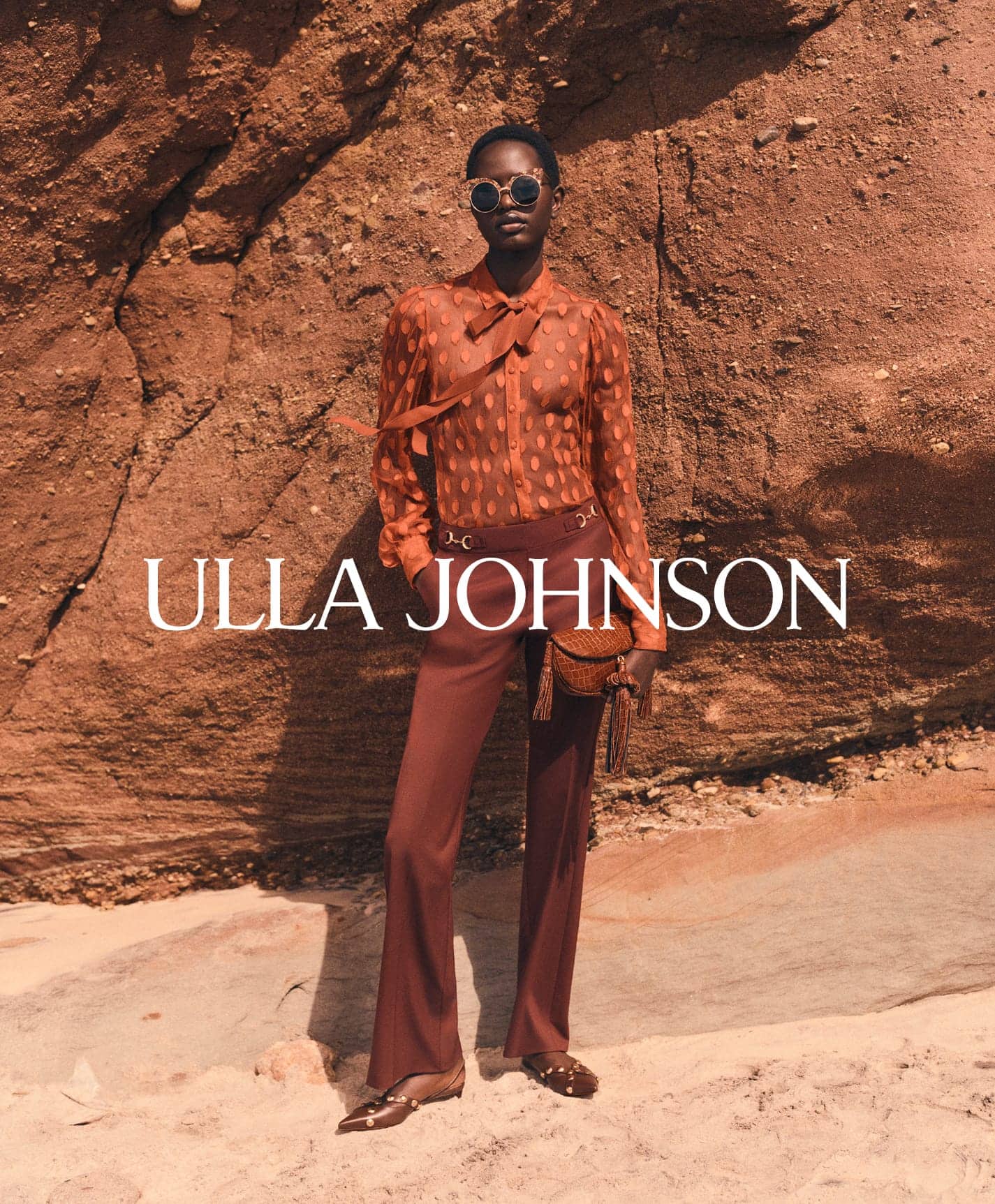 Model wearing sheer orange blouse and brown trousers standing against a rocky cliffside background.