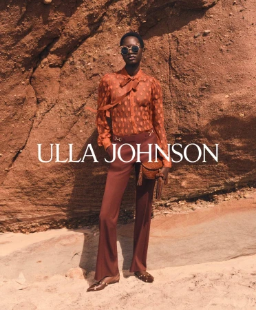 Model wearing sheer orange blouse and brown trousers standing against a rocky cliffside background.