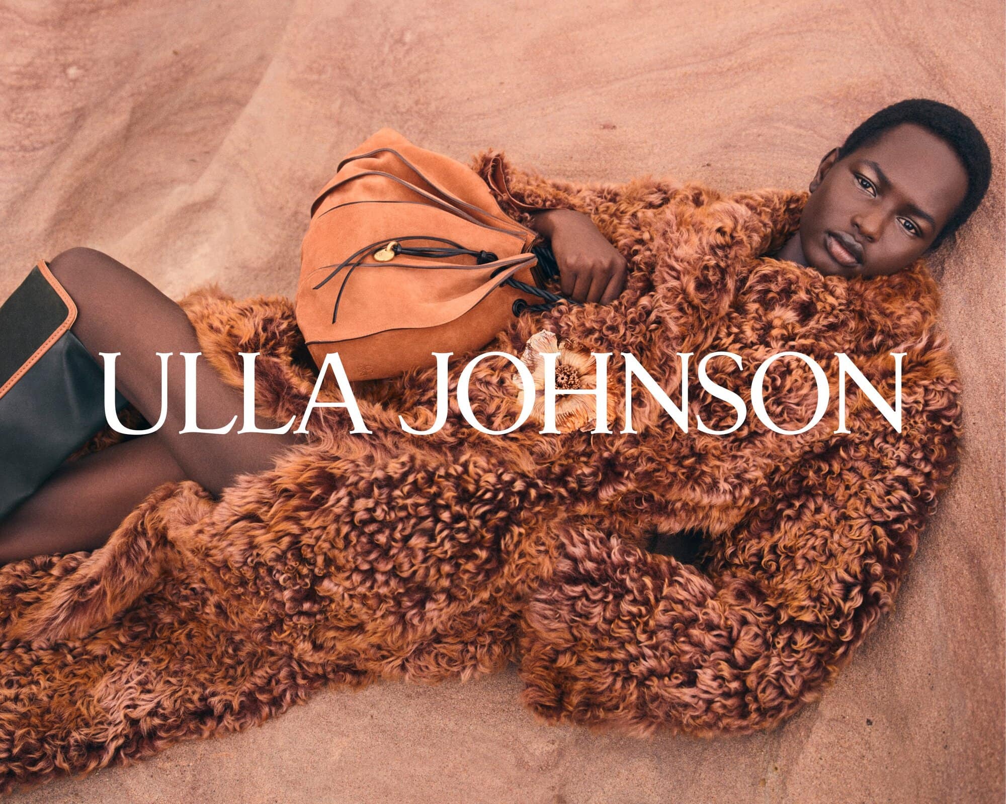 Model in textured brown shearling coat reclining on sand dunes next to a suede bag with brand text overlaid