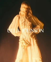 Blonde model in a white ruffled tiered dress with voluminous long sleeves holding a gold pouch bag against a dark background