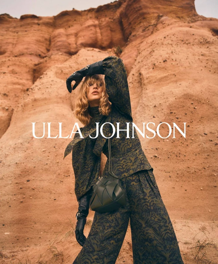 Model posing against a rugged earthen rock cliff wearing a patterned dark green top and matching trousers with leather gloves.