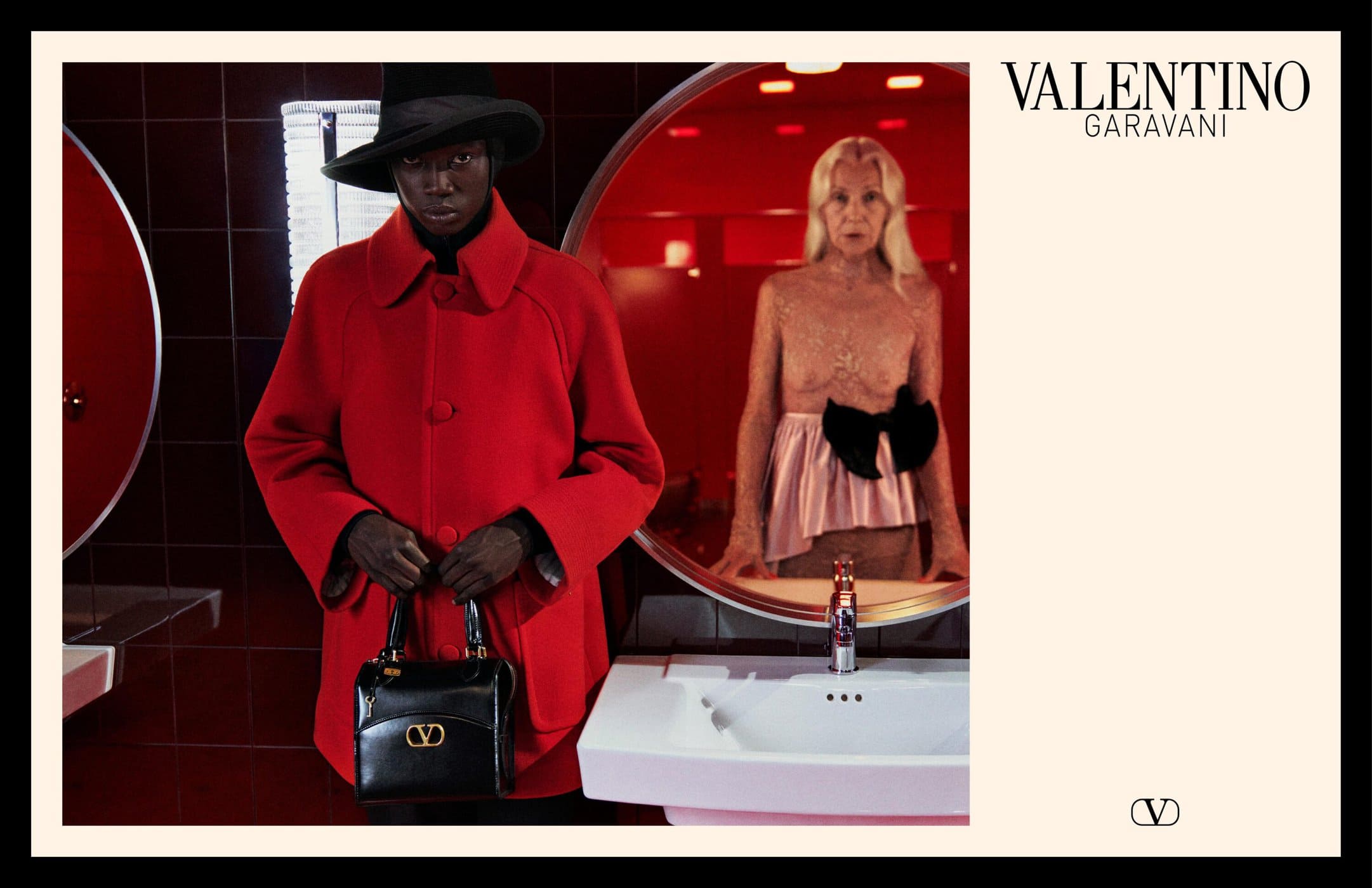 Model wearing a vibrant red coat and black wide-brim hat holding a black handbag in a tiled interior with a round mirror reflection