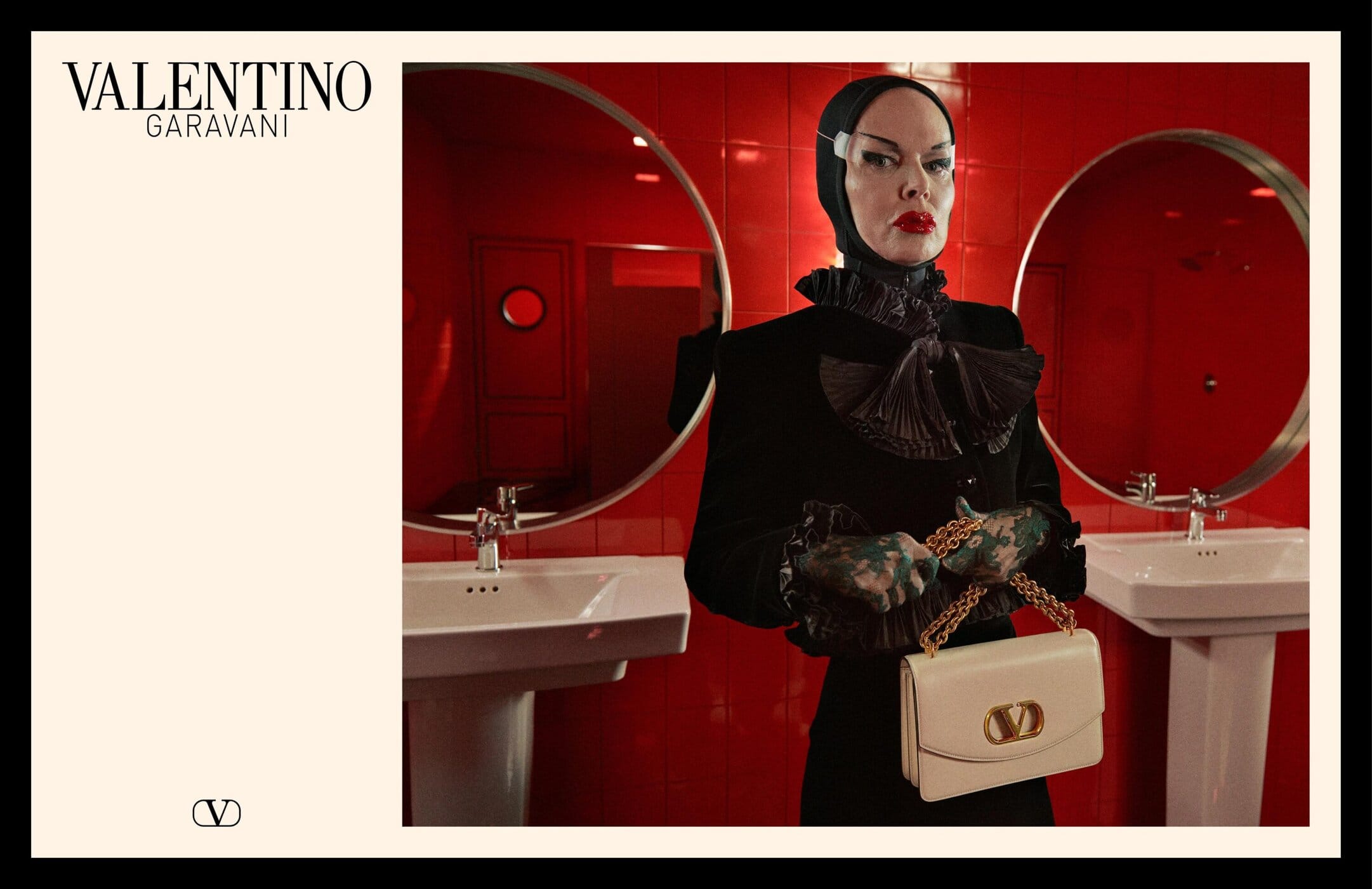 Model in dark jacket with ruffled collar holding a white chain handbag in a red tiled restroom with round mirrors
