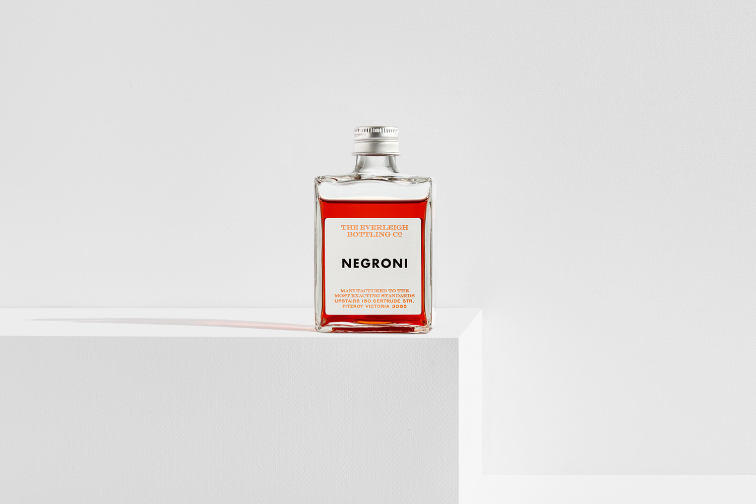 A rectangular glass cocktail bottle containing red liquid sitting on a white pedestal against a plain white background.