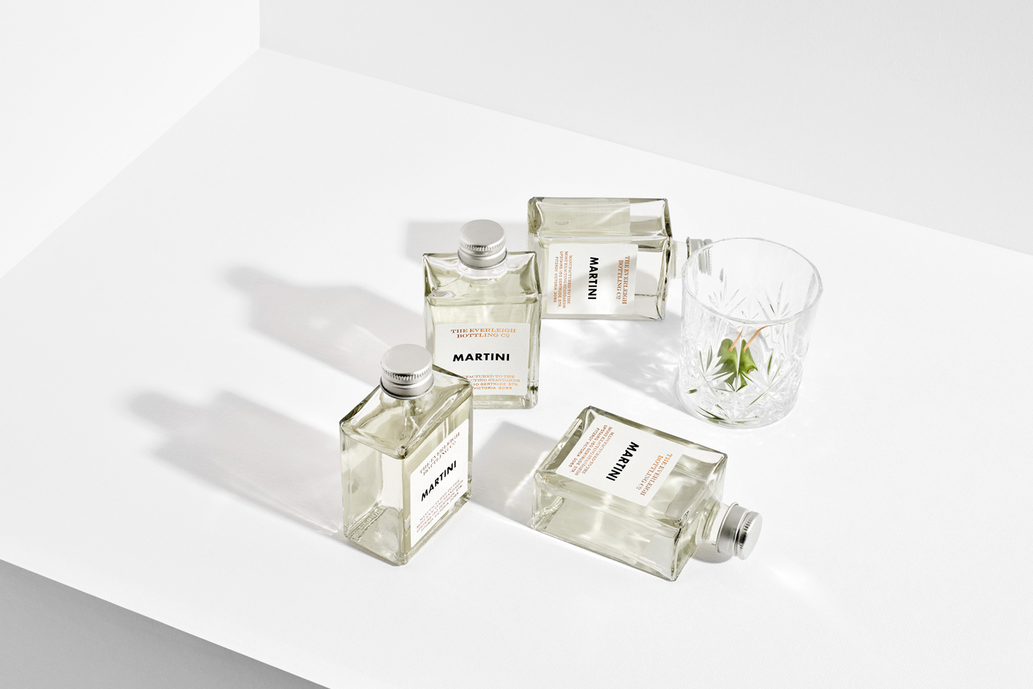 Four square glass bottles of martini and a crystal glass on a clean white surface with sharp shadows