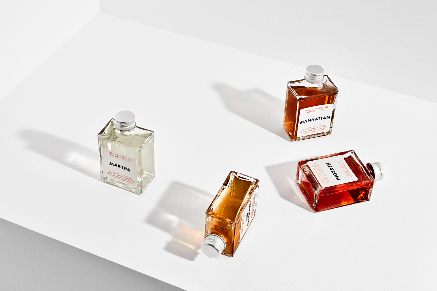 Four square glass bottles of cocktail beverages arranged on a plain white surface with crisp shadows.