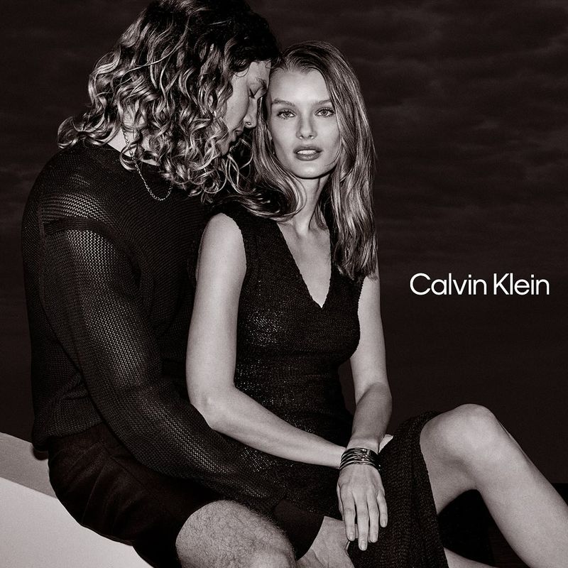 Monochrome image featuring a woman in a black dress seated with a man; the Calvin Klein logo is visible on the right.