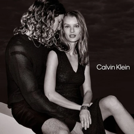 Monochrome image featuring a woman in a black dress seated with a man; the Calvin Klein logo is visible on the right.