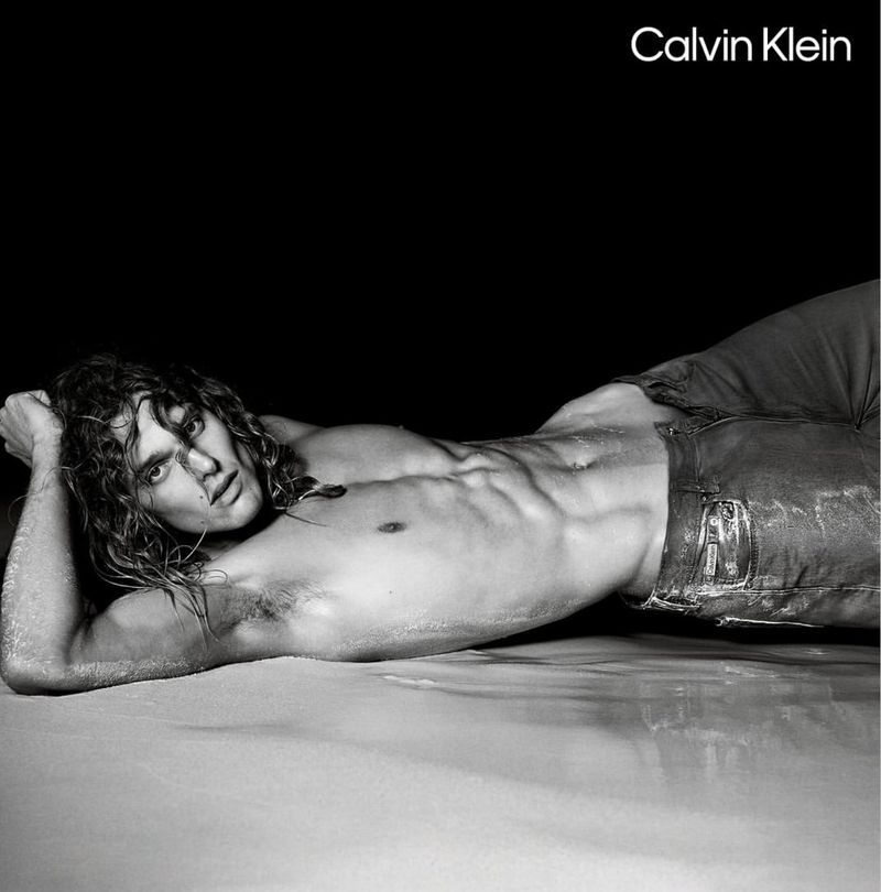 Shirtless adult male model with long wavy hair poses while laying on sand, wearing jeans, lit with bright studio lighting, and a black background.