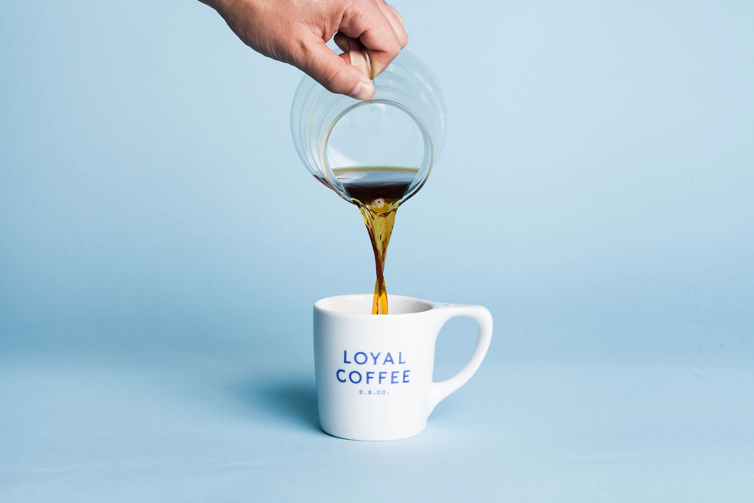 A hand pours coffee from a glass carafe into a white ceramic mug against a light blue seamless background.