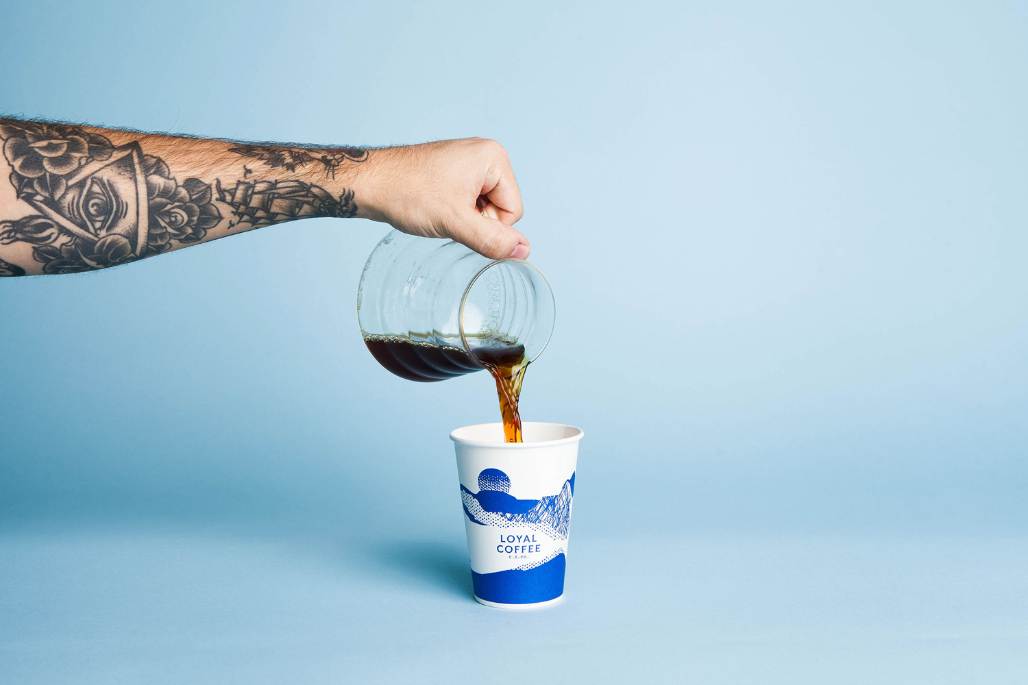 Tattooed arm pouring coffee from a glass carafe into a paper cup against a light blue background