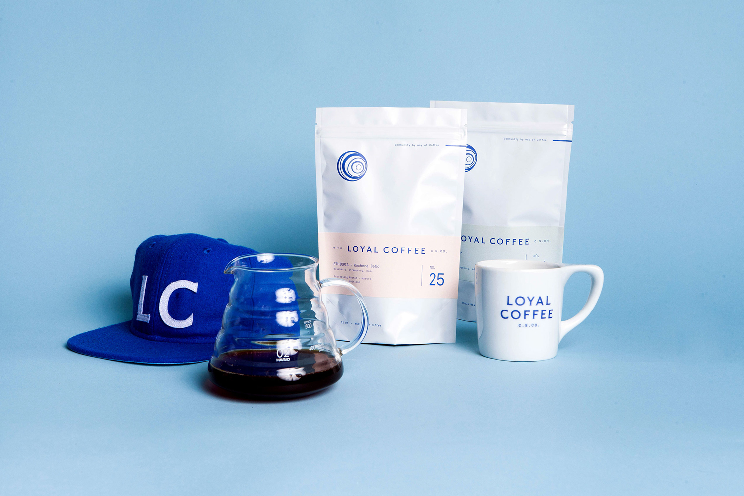 Loyal Coffee brand identity display featuring two coffee bags, a glass server, a mug, and a blue cap on a blue background