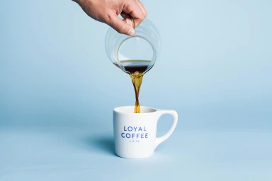 A hand pours coffee from a glass carafe into a white ceramic mug against a light blue seamless background.