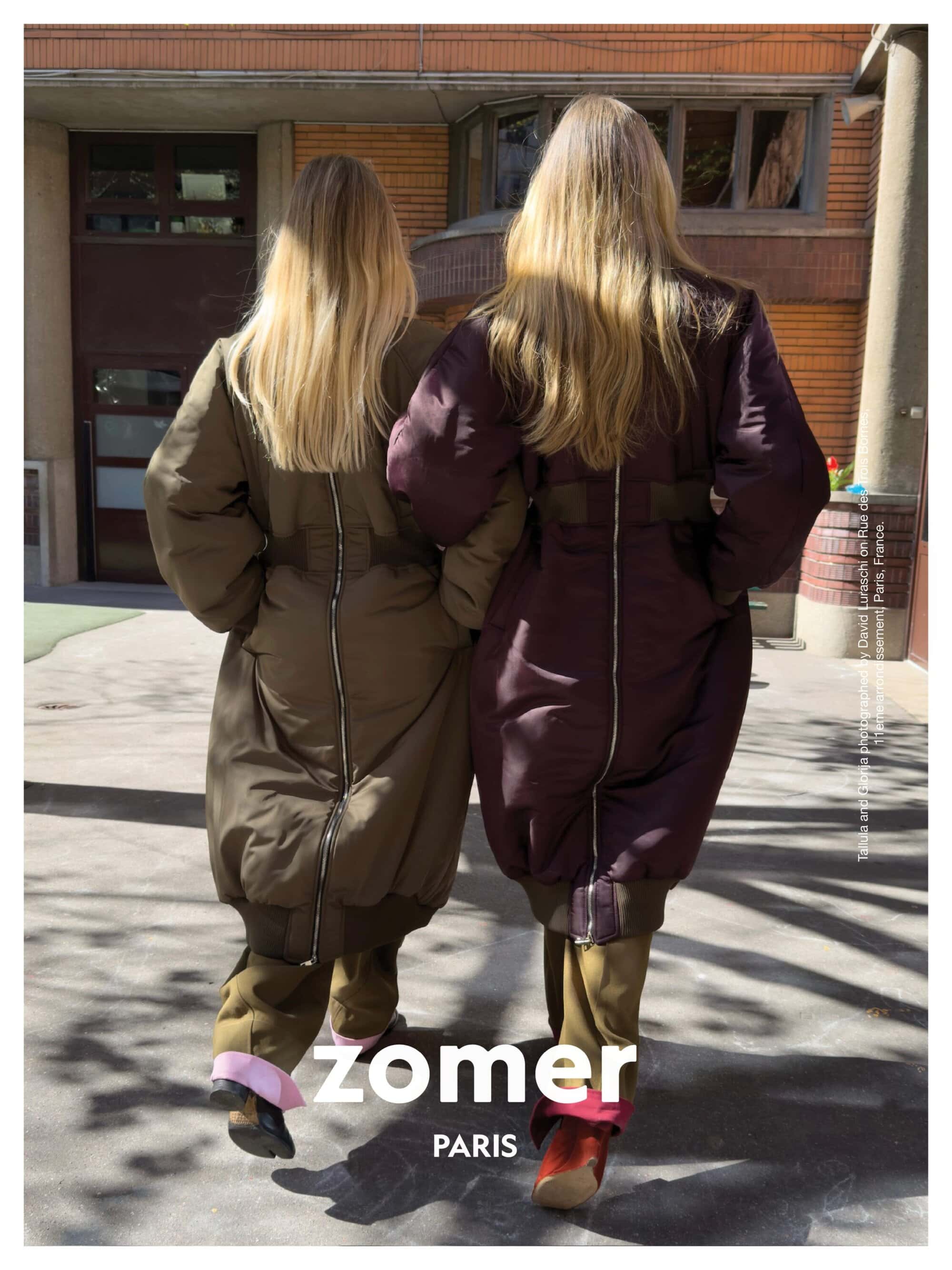Two models seen from behind wearing long padded coats with back zippers outside a building for Zomer campaign