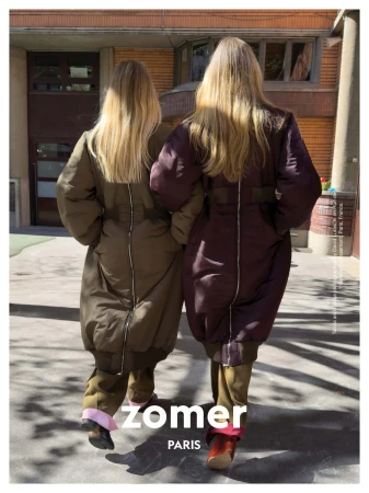 Two models seen from behind wearing long padded coats with back zippers outside a building for Zomer campaign